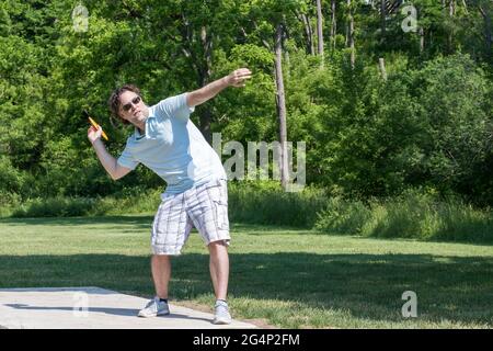 Uomo caucasico di media età in shorts, camicia da golf, e occhiali da sole, preparandosi a lanciare un disco giallo nel gioco del disc golf. Allineando il suo colpo. Foto Stock