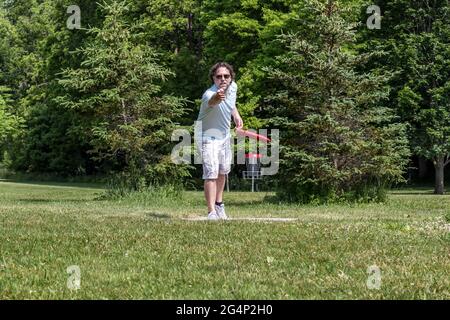 Uomo caucasico di media età in maglietta, pantaloncini corti e occhiali da sole che giocano a disc golf. Testa sulla vista di lui che lancia il disco con il cestino da golf disco sullo sfondo. Foto Stock