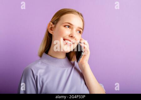 Giovane bella donna che parla tramite cellulare smartphone isolato su sfondo viola colore. Felice sorridente caucasica adolescente ragazza uso cellulare Foto Stock