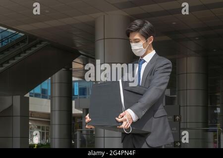 Uomo asiatico dipendente con maschera lasciando ufficio con casella a causa di licenziamento, covid 19 effetti nel business Foto Stock