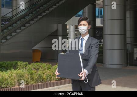 Uomo asiatico dipendente con maschera lasciando ufficio con casella a causa di licenziamento, covid 19 effetti nel business Foto Stock