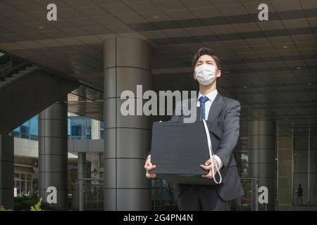 Uomo asiatico dipendente con maschera lasciando ufficio con casella a causa di licenziamento, covid 19 effetti nel business Foto Stock