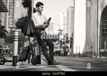 giovane uomo d'affari asiatico in piedi accanto al suo scooter elettrico sulla strada in giù di città moderna, in bianco e nero Foto Stock