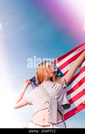 Giovane bionda millenaria in piedi con bandiera americana accanto al cielo blu. Bandiera degli stati uniti nelle sue mani. 4 luglio giorno dell'Indipendenza. Patriottismo USA festa nazionale. Caucasica ragazza. Spazio di copia. Libertà e concetto memoriale Foto Stock