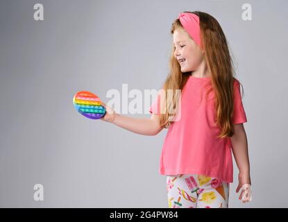 Felice ragazza di capretto sorridente in vestiti rosa tiene e guarda il nuovo giocattolo rotondo di colore di arcobaleno sensoriale - schioccarlo Foto Stock