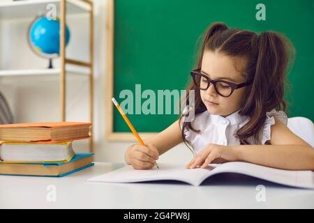 Concetto di ritorno a scuola. Una ragazza intelligente in occhiali scrive con una matita in un notebook mentre si siede a un tavolo in classe. Foto Stock