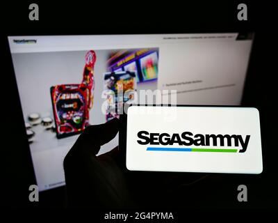 Persona che tiene il cellulare con il logo della società di gioco giapponese sega Sammy Holdings Inc. Sullo schermo davanti alla pagina web. Mettere a fuoco il display del telefono. Foto Stock