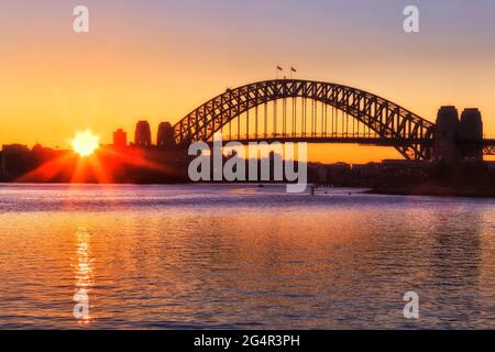 Il sole sorge sull'orizzonte dietro il ponte del porto di Sydney che splende il cielo australiano di colore giallo e rosso - le acque calde del porto di Sydney. Foto Stock