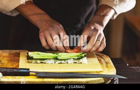 Cuocete le mani per preparare il sushi giapponese. Il processo di cottura del sushi roll con salmone, cetriolo, riso e nori. Foto Stock