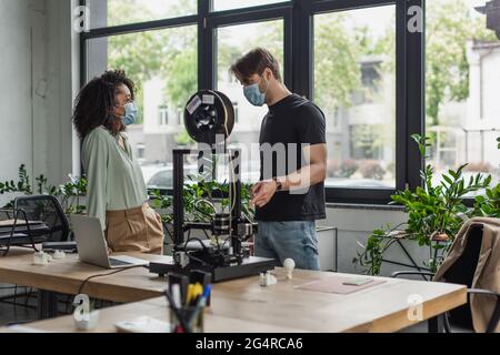 Progettisti interrazziali in maschere mediche che parlano vicino stampante 3D in ufficio moderno Foto Stock