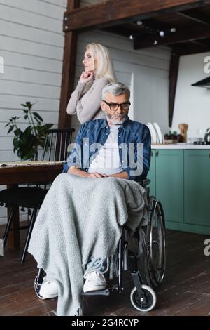 Triste uomo disabile in sedia a rotelle che guarda lontano vicino moglie a casa Foto Stock