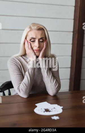Triste donna anziana con demenza seduta vicino al seghetto alternativo offuscato sul tavolo Foto Stock