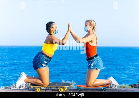 Due amici femminili con maschere protettive che giocano con skateboard. Girlfriends festeggiando il loro successo, l'alta amicizia e il coronavirus concep Foto Stock