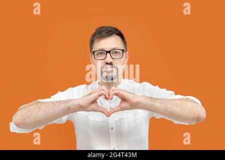 Ritratto di bell'uomo sorridente in camicia bianca che mostra il segno del cuore isolato su sfondo arancione. Foto Stock