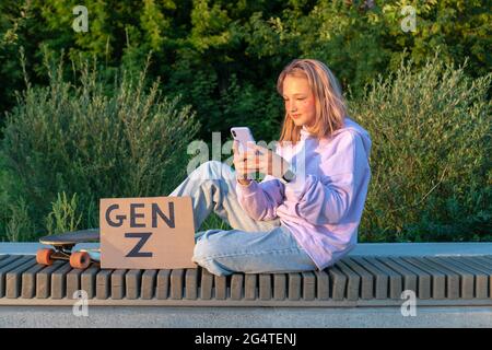 Un'elegante ragazza adolescente in un jeans lilla con felpa con cappuccio e capelli rosa si siede su una panca con un telefono in mano accanto a una tavola lunga e un segno con l'iscrizione Foto Stock