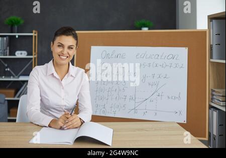 Ritratto di felice giovane insegnante seduto alla scrivania durante la sua classe di matematica online Foto Stock