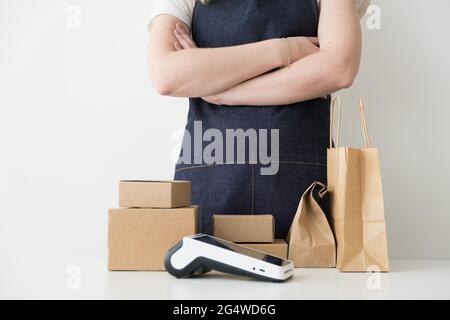 Donna che indossa un grembiule in denim con le braccia incrociate in piedi dal tavolo con scatole di cartone, confezioni di cartone e moderno terminale POS di pagamento. Piccolo autobus Foto Stock