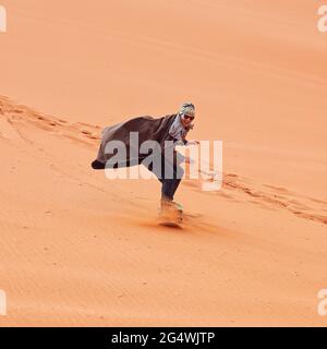 Giovane uomo sorridente, sabbia duna surf con bisht - cappotto beduino tradizionale. Il Sandsurfing è una delle attrazioni del deserto di Wadi Rum Foto Stock