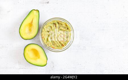 Due metà di avocado, e una ciotola di vetro con guacamole preparato su una scrivania in pietra bianca, vista dall'alto per il testo sul lato destro Foto Stock
