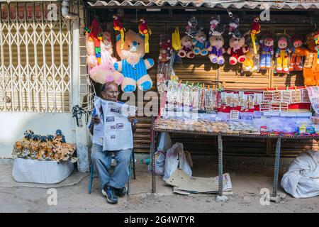 Il maschio indiano baldante siede il giornale di lettura mentre la stalla della strada che mangia vende i giocattoli coccolati e i monili dell'oro, Udupi, Karnataka, India Foto Stock