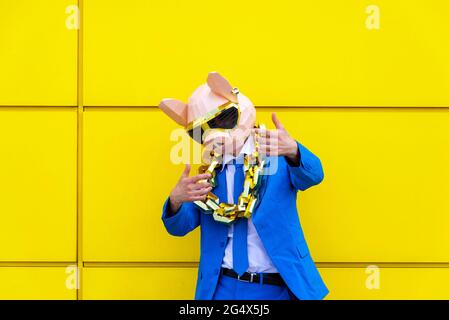 Uomo che indossa una vivace tuta blu, una maschera di maiale e una grande catena dorata che si posano contro la parete gialla Foto Stock