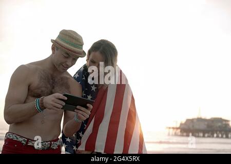 Bell'uomo che usa il tablet digitale della ragazza avvolto in bandiera sulla spiaggia di Santa Monica Foto Stock