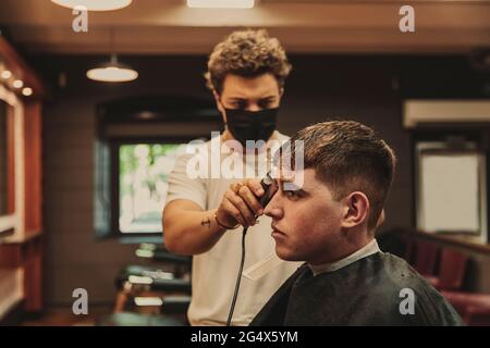 Giovane uomo che ottiene il taglio dei capelli da barbiere maschio in salone durante l'epidemia di COVID-19 Foto Stock