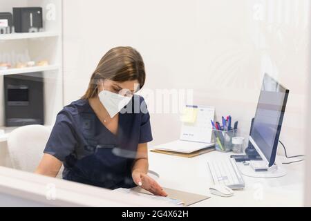 Dentista femminile che indossa una maschera protettiva che lavora alla scrivania in clinica Foto Stock