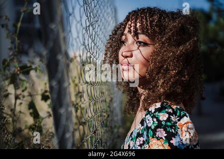 Giovane donna con capelli ricci in piedi da recinzione che guarda via Foto Stock