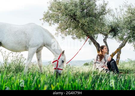 Giovane donna seduta di fronte all'albero mentre il cavallo pascolava sul campo Foto Stock