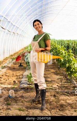 Sorridente agricoltore femmina con annaffiatura lattina e attrezzi a mano in piedi a serra Foto Stock