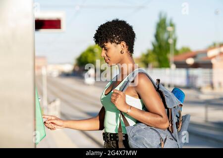 Giovane donna che trasporta lo zaino mentre usa la macchinetta del biglietto Foto Stock