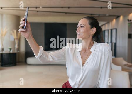 Donna d'affari sorridente che prende selfie attraverso lo smartphone nella lobby Foto Stock