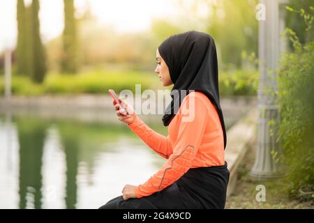 Giovane donna che usa lo smartphone mentre si siede vicino al laghetto Foto Stock