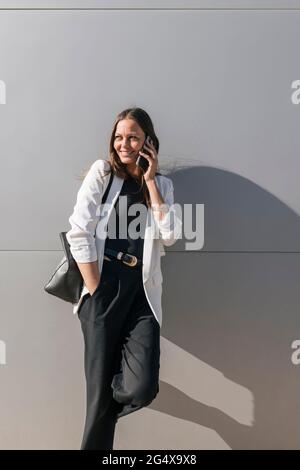 Donna d'affari sorridente con la mano in tasca che parla sul telefono cellulare di fronte alla parete Foto Stock