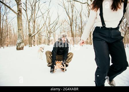 Donna che tira l'uomo seduto su una slitta di legno con il cane dietro in foresta Foto Stock
