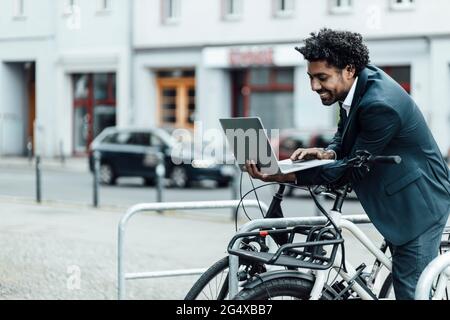 Uomo d'affari sorridente che usa il computer portatile mentre si appoggia in bicicletta al parcheggio della città Foto Stock