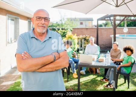 Uomo anziano in piedi con le braccia incrociate mentre la famiglia in background in cortile Foto Stock