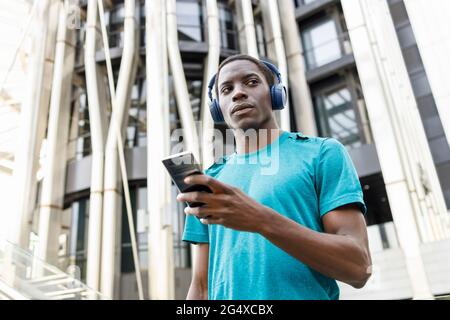 Uomo che indossa cuffie wireless che tengono il telefono cellulare davanti all'edificio Foto Stock