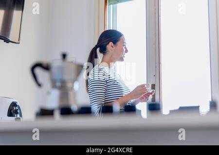 Giovane donna che guarda attraverso la finestra mentre tiene la tazza di caffè in cucina Foto Stock
