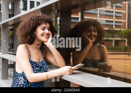 Giovane donna con la mano sul mento e le cuffie intrauricolari sorridono mentre si tiene il telefono cellulare Foto Stock