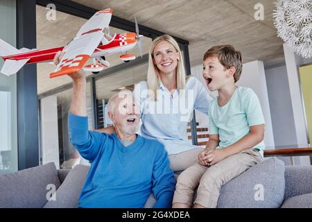 Uomo anziano che tiene un aeroplano giocattolo che gioca con il nipote mentre si siede da donna in soggiorno Foto Stock