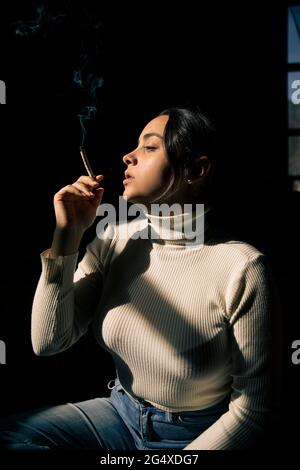 Giovane donna che fuma la sigaretta alla luce del sole a casa Foto Stock