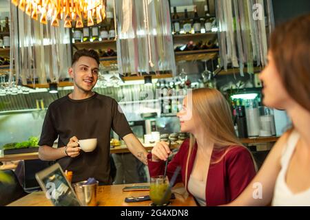 Un giovane sorridente in piedi con una tazza di caffè che parla con le amiche seduti al tavolo al bar Foto Stock