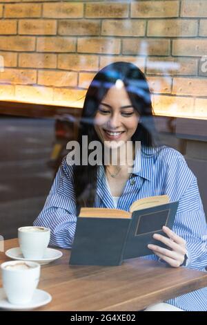 Sorridente giovane donna che legge libro mentre si siede in bar visto attraverso il vetro Foto Stock