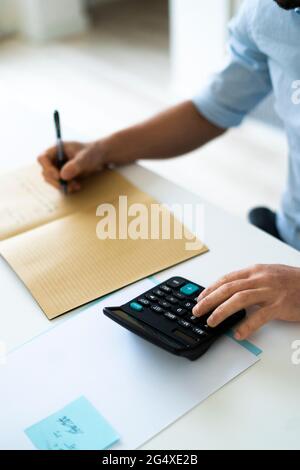 Uomo d'affari che scrive nel libro mentre usando la calcolatrice in ufficio Foto Stock