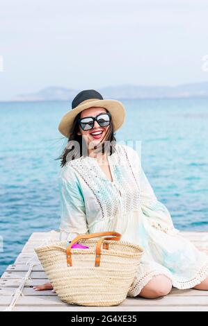 Donna matura felice che indossa cappello e occhiali da sole seduti sul molo Foto Stock