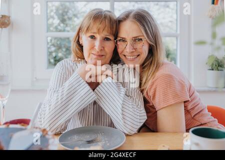 Figlia sorridente abbracciando la madre mentre si siede al tavolo da pranzo Foto Stock