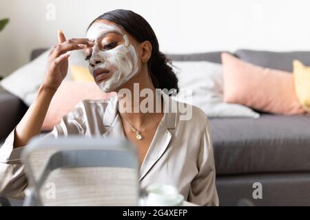 Giovane donna che guarda nello specchio mentre applica il prodotto di bellezza a casa Foto Stock