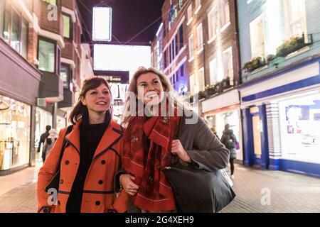 Sorridendo le amiche che indossano cappotti invernali che guardano via mentre camminano insieme in città durante la notte Foto Stock